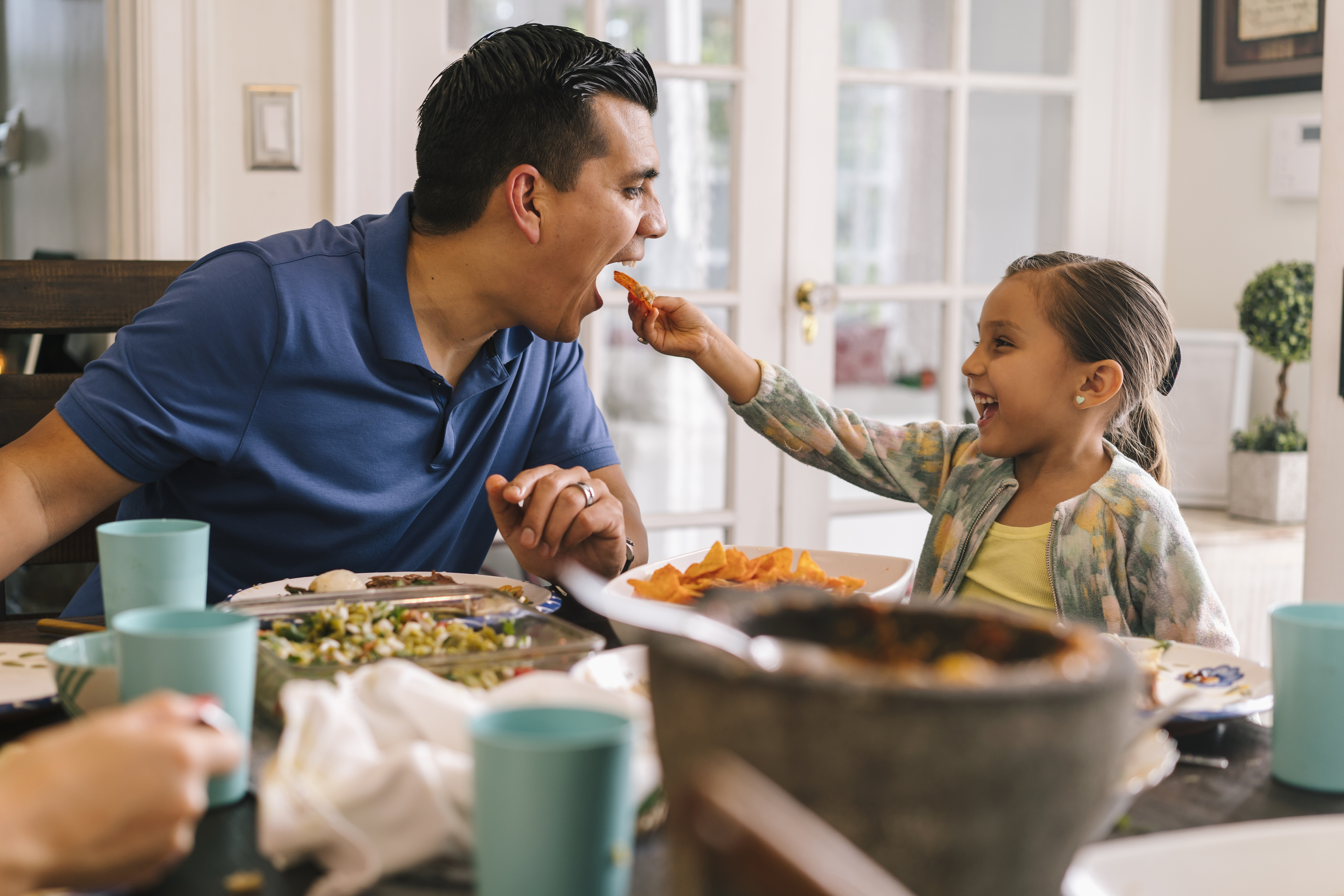 Padre e hija latinos sonriendo en una mesa de comedor mientras ella le da de comer una papa frita de manera juguetona.